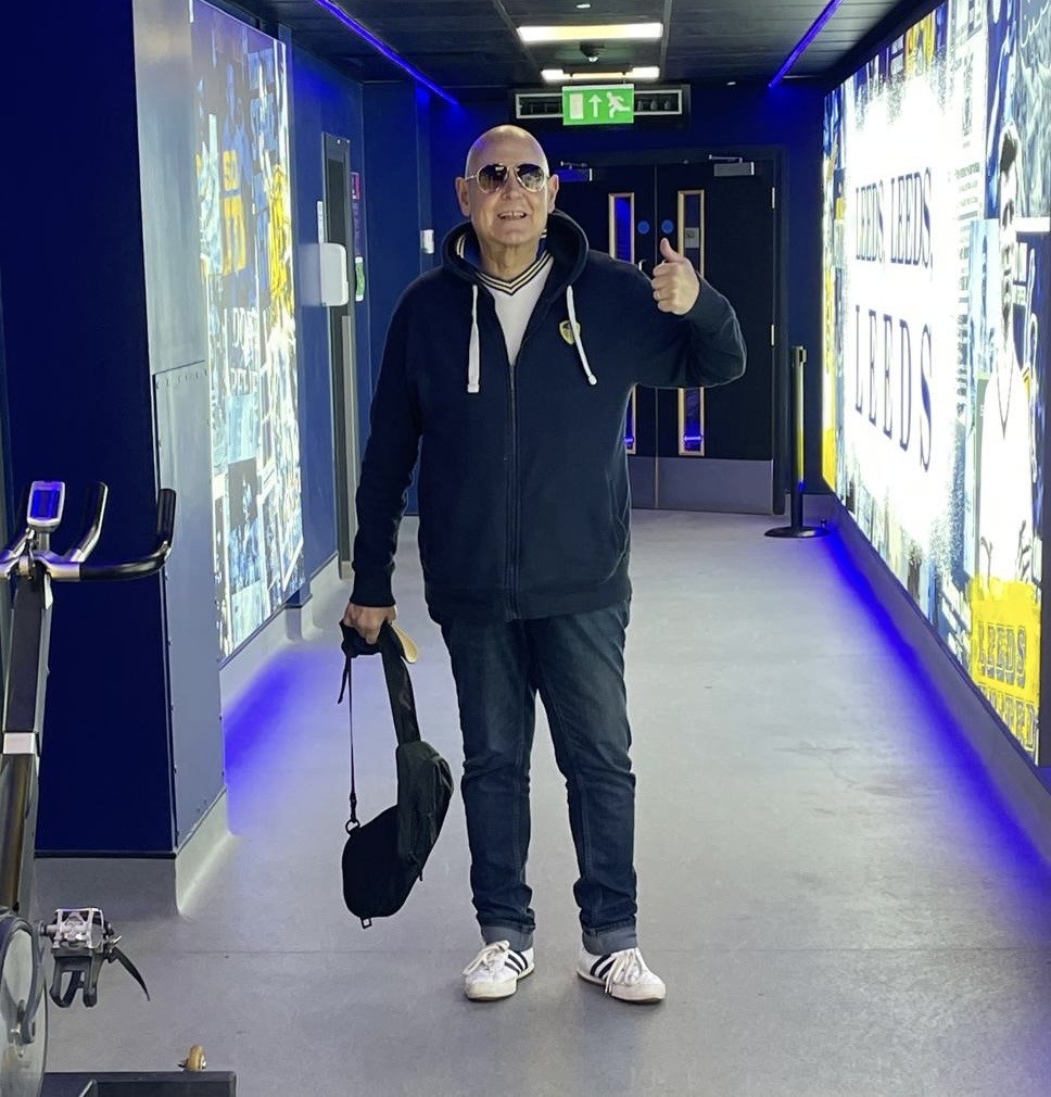 An image of a person giving the thumbs up in a corridor with Leeds United Football Club imagery and symbolism on the walls behind him to his left and right,